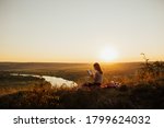 Young woman reading book at sunset in front of beautiful landscape with fields and river.  Beautiful girl sitting on grass and reading a book.