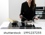 Cropped shot of woman boiling water in metal pot to cook spaghetti, holding glass lid and big spoon while cooking on black ceramic induction cooktop in white minimalistic kitchen