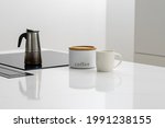 Brewing coffee for breakfast in steel pot on built in induction ceramic cooktop, white labeled jar containing coffee with mug next to it in kitchen with minimalistic design