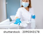 Nurse in protective gloves cleaning work surface at stomatology clinic, sanitizing table with disinfectant spray bottle, washing dental chair with towel, selective focus