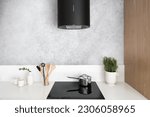 stylish kitchen with induction glass ceramic stove, modern hood extractor, kitchenware, cups and houseplant on white countertop