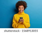 Small photo of Adorable dark skinned adult woman dressed in yellow jumper, holds mobile phone, browses social networks, has broad smile, enjoys online chatting, isolated on blue wall, being always in touch