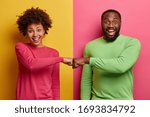 Small photo of Happy dark skinned ethnic woman and man make fist bumps, work as team, agree to do something, smile positively, pose against bright two colored background. Partnership and collaboration concept