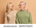 Small photo of Caring middle aged woman embraces her husband looks with love and broad smile. Married mature couple have good relationships isolated on brown background. Smiling aged husband and wife pose for photo