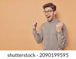 Small photo of Overjoyed positive man clenches fists with triumph rejoices excellent news exclaims from happiness cannot believe in success wears casual jumper and spectacles gesture actively poses indoor.