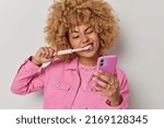 Small photo of Dental care concept. Young European woman with curly hair cleans teeth with electric toothbrush undergoes daily hygiene routines in morning scrolls newsfeed via smartphone wears pink jacket.