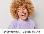 Small photo of Portrait of happy positive woman keeps eyes closed smiles broadly shows white teeth dressed in purple pullover enjoys life feels very glad isolated over grey studio background. People and emotions