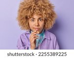 Small photo of Close up shot of beautiful woman with curly hair concentrated with shocked scrupulous gaze at camera keeps hand on chin reacts on something terrifying dressed in jacket stands over purple background