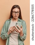 Small photo of Stunned brunette woman scrolls newsfeed on smartphone stares wondered at smartphone screen wears transparent eyeglasses and casual shirt isolated over brown background. People and technology