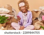 Small photo of Happy curly haired woman checks newsfeed on smartphone sits crossed legs on floor among cardboard boxes prepares for relocation wears casual clothing applies beauty patches beige background.