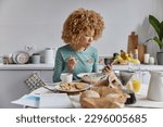 Small photo of Indoor shot of curly haired young woman checks social media and newsfeed while having breakfast poses at kitchen eats cereals and drinks coffee undergoes beauty procedures focused at smartphone
