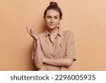 Small photo of Waist up shot of confused European woman shrugs shoulders feels unaware wears brown shirt cannot understand what to do isolated over beige background. Peope reactions and perception concept.