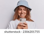 Small photo of Curious young pretty woman in denim panama and pullover bites lips concentrated in smartphone screen reads something interesting and intriguing poses against white background watches video online