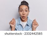 Small photo of Portrait of dark haired Latin woman holds breath keeps mouth opened hands clenched in fists startled by unsettling news got unexpected offer dressed in denim clothing isolated over white background