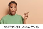 Small photo of Horizontal shot of African guy gazes wide eyed at camera his jaw dropped in absolute shock witnesses something truly astonishing points index finger on blank space poses over brown background