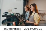 Small photo of Beautiful young Asia lady barista working with coffee machine in coffee shop. Two small business owner Korean girl in apron making coffee by coffee machine with friend at counter in urban cafe.