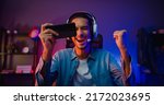 Happy asia man gamer wear headphone competition play video game online with smartphone colorful neon light in living room at night modern house. Esport streaming game online, Home quarantine activity.