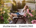 Young romantic woman holding book to her chest on urban rooftop garden at sunset. Dreamy girl reading exciting novel while sitting in chaise longue on cozy terrace with flowers, plants and city view.