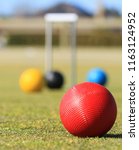 Small photo of In the foreground is a red croquet ball on a green croquet lawn in Australia with a defocused croquet hoop and balls in the background