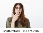 Mom woke up daughter very early. Shot of sleepy attractive caucasian woman with messy brown hair, wearing glasses, feeling tired after night without sleep, yawning, covering opened mouth with palm