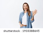 Small photo of Cheerful friendly girl saying hello, winking and smiling, waving hand at camera, say hi and greet you, standing against white background