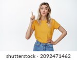 Small photo of Cute blond girl pucker lips, showing something up, pointing finger at advertisement or sale banner, standing in yellow t-shirt and jeans, white background