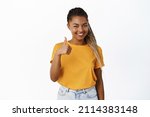 Small photo of Very good. Smiling satisfied Black girl shows thumbs up and nod in approval, like something, standing in yellow t-shirt over white background