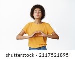 Small photo of Romantic cute young african american woman pucker lips, reaching for kiss, showing I love you heart hand sign, standing in yellow tshirt over white background