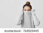 Portrait of girl pulling her trendy sweater over head having fun. Woman with tied hair in topknot being childish disappearing in her clothes looking from underneath. Happiness concept
