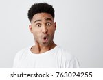 Close up of funny handsome young dark-skinned man with afro haircut in stylish white t-shirt looking in camera with raised eyebrows and surprised face expression.