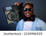 Stylish smiling black man wears black glasses and denim shirt. Popular DJ with an earring in his ear and headphones carries DJ console on his shoulder to concert to entertain audience with music track