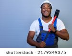 Small photo of African-American man in a white helmet and work clothes smiles with a toothy smile, holding a sledgehammer in his hands on a gray background. Portrait of a happy worker in overalls with a copy space