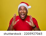 Shocked young African-American Santa Claus in a red T-shirt and a Christmas hat, shouting with a hand gesture at his mouth, isolated on a yellow background. Concept Happy New Year, Merry Christmas