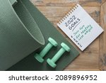 Notepad on the wooden background with the goals for 2022 - join gym, eat healthy, loose weight. Green dumbbells and sports mat