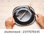 Small photo of On a black plate, an ear, in the hands of a fork and a knife. A symbol of hunger and inept housekeeping is a plate with cutlery and a spikelet against the background of a wooden structure.