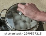 Small photo of Pot of boiling water in which white chicken eggs are boiled. Water boils and boils in pan with boiled eggs. Female hand removes transparent lid from pan with boiled white chicken eggs. Close-up.