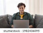 Young cheerful man sitting on sofa with laptop indoors looking at computer monitor and smiling. Handsome young man enjoying watching funny video on social media