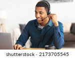 Small photo of African american young man customer support call center operator or receptionist sitting at the workplace in a modern office consulting a client, uses a headset, smiles friendly