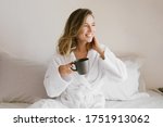 Young beautiful woman wearing white bathrobe having breakfast in bed with coffee and croissant and fresh fruits in cozy bedroom. Morning rituals.