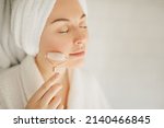 Beautiful young woman with perfect skin wearing white bathrobe and towel on head after shower, making face massage using a jade face roller with natural quartz stone in bathroom.