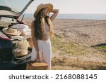 Young beautiful woman standing near car with summer attributes, enjoying sunset on the beach. Summer vacation.
