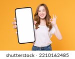 Cheerful beautiful Asian woman holding smartphone and shows ok sign on light yellow background.