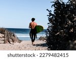 Surfer with wetsuit getting ready for a surf session. Boy walking on the beach with a green surfboard under the arm. Back point of view. Ocean waves and surf. Surfing in Australia.