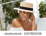 Pretty glad female wears straw hat, shows her tanned bare shoulder, smiles with happy expression as looks joyfully aside, spends summer vacations abroad. Lovely young woman enjoys good resort alone