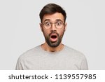Small photo of Stunned emotive bearded man keeps mouth widely opened, looks with bated breath, hears terrific news, wears spectacles, isolated over white studio wall. Surprised hipster sees something unbelievable