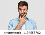 Image of hesitant unshaven European male with thick beard, holds chin, purses lips with clueless expressions, doubts what to eat delicious for supper, isolated over white background. What to choose?