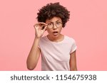 Small photo of Horizontal shot of attractive African American female with scrupulous expression, looks with indignation through eyewear, opens mouth, tries to read something from distance, poses indoor alone