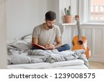Photo of serious man with dark bristle, writes down story in diary about what happened during day, sits crossed legs at bed, wears casual clothing, has day off or weekend. Male records information