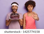 Say no to harmful eating! Serious dark skinned woman shows stop gesture, stands near man who holds big tasty cake with much calories, asks not to eat such food if you want to be in good shape