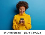 Photo of dark skinned lady enjoys distant communication, uses mobile phone, surfes fast unlimited internet, has pleasant smile, dressed in yellow sweater, makes shopping online, isolated on blue